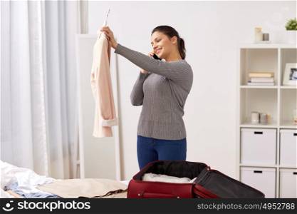 tourism, business trip, people and luggage concept - happy young woman calling on smartphone and packing travel bag at home or hotel room. woman with smartphone packing travel bag at home