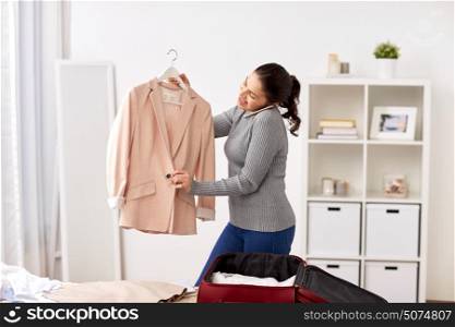 tourism, business trip, people and luggage concept - happy young woman calling on smartphone and packing travel bag at home or hotel room. woman with smartphone packing travel bag at home
