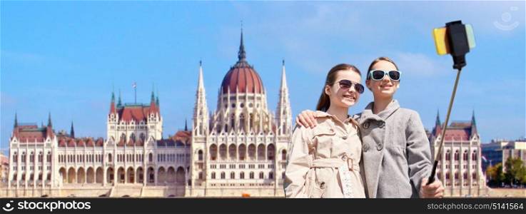 tourism, childhood and travel concept - happy girls taking picture with smartphone on selfie stick over house of parliament in budapest. girls with smartphone selfie stick in budapest