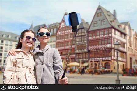 tourism, childhood and travel concept - happy girls taking picture with smartphone on selfie stick over frankfurt am main city street background. girls with smartphone selfie stick in frankfurt