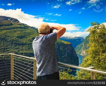 Tourism holidays picture and traveling. Mature male tourist enjoying fjord landscape Geirangerfjord from Ornesvingen viewpoint, taking photo with camera, Norway Scandinavia.. Tourist taking photo of fjord landscape, Norway