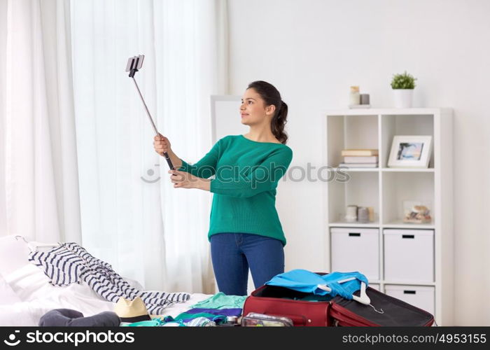 tourism, people and luggage concept - happy young woman packing travel bag at home or hotel room and taking picture with smartphone selfie stick. woman packing travel bag at home or hotel room