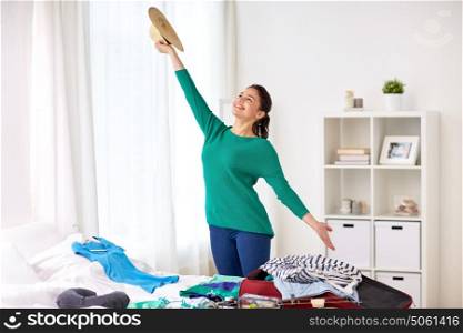 tourism, people and luggage concept - happy young woman with hat packing travel bag at home or hotel room. woman packing travel bag at home or hotel room