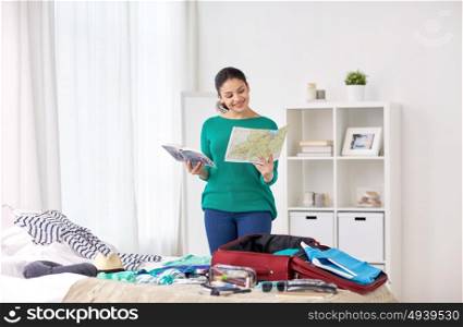 tourism, people and luggage concept - happy young woman with map and city guide packing travel bag at home or hotel room. woman packing travel bag at home or hotel room