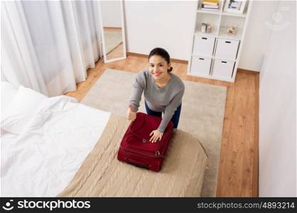 tourism, people, gesture and luggage concept - happy young woman packing travel bag at home or hotel room and showing thumbs up