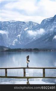 Tourism theme image about a woman with a photo camera, walking on a wooden deck over the Hallstatter lake, surrounded by Northern Limestone Alps, Austria.