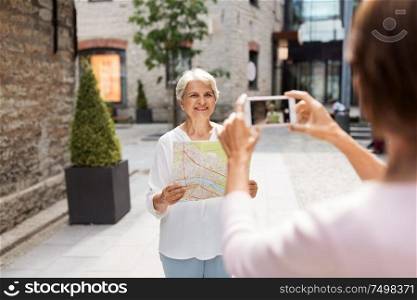 tourism, travel and friendship concept - happy senior woman with city map being photographed by her friend on street in tallinn. senior women with city map on street in tallinn