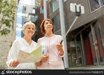 tourism, travel and friendship concept - happy senior women with map and city guide taking picture by smartphone on selfie stick on street in tallinn. senior women with map and city guide taking selfie