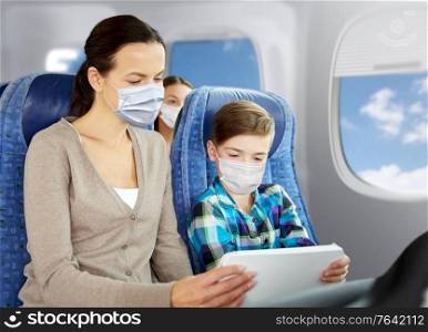 tourism, travel and pandemic concept - happy mother and son wearing face protective medical masks for protection from virus disease with tablet pc computer sitting in plane over porthole background. mother and son in masks traveling by plane