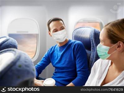 tourism, travel and pandemic concept - happy passengers or tourists wearing face protective medical masks for protection from virus disease with coffee talking in plane over porthole background. couple of passengers in masks travelling by plane