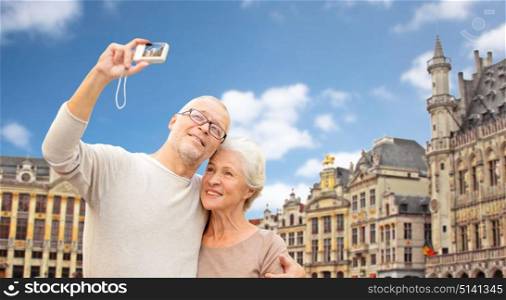 tourism, travel and people concept - happy senior couple with camera taking selfie over grand place in brussels city background. senior couple with camera traveling in brussels