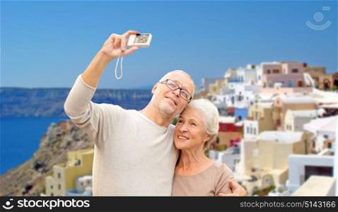 tourism, travel and people concept - happy senior couple with camera taking selfie over santorini island background. senior couple with camera travelling in santorini