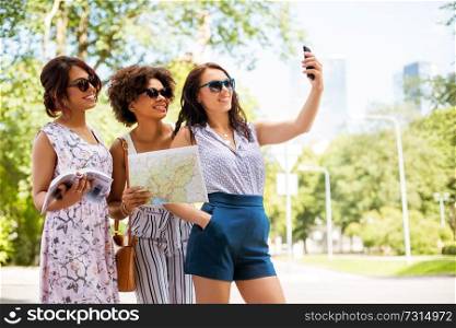 tourism, travel and technology concept - happy women with city guide and map taking selfie by smartphone in summer park. women with city guide and map taking selfie