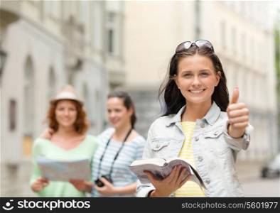 tourism, travel, leisure, holidays and friendship concept - smiling teenage girls with city guide, map and camera showing thumbs up outdoors