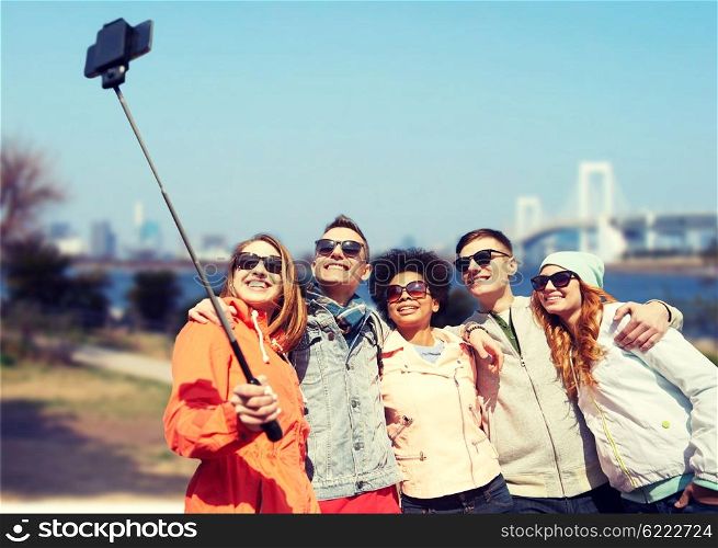tourism, travel, people, leisure and technology concept - group of smiling teenage friends taking selfie with smartphone and monopod over rainbow bridge in tokyo city