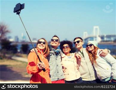 tourism, travel, people, leisure and technology concept - group of smiling teenage friends taking selfie with smartphone and monopod over rainbow bridge in tokyo city