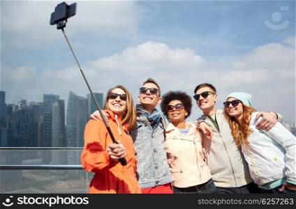 tourism, travel, people, leisure and technology concept - group of smiling teenage friends taking selfie with smartphone and monopod over singapore city background