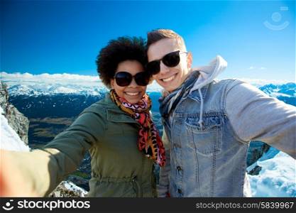 tourism, travel, people, leisure and technology concept - happy international teenage couple taking selfie over alps mountains in austria background