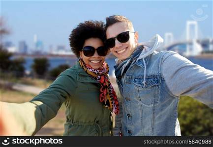 tourism, travel, people, leisure and technology concept - happy international teenage couple taking selfie over rainbow bridge at tokyo in japan background