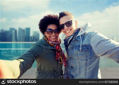 tourism, travel, people, leisure and technology concept - happy international teenage couple taking selfie over singapore city background