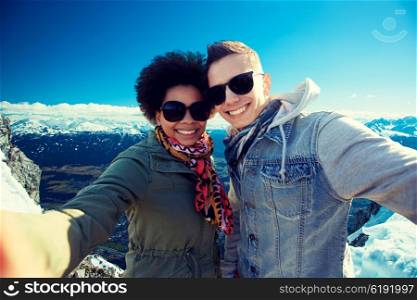 tourism, travel, people, leisure and technology concept - happy international teenage couple taking selfie over alps mountains in austria background