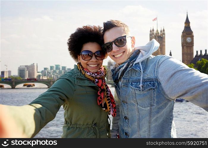 tourism, travel, people, leisure and technology concept - happy teenage international couple taking selfie over houses of parliament and thames river in london background