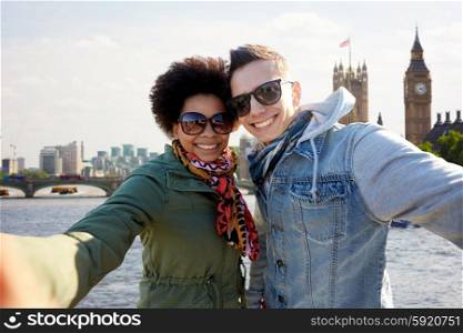 tourism, travel, people, leisure and technology concept - happy teenage international couple taking selfie over houses of parliament and thames river in london background