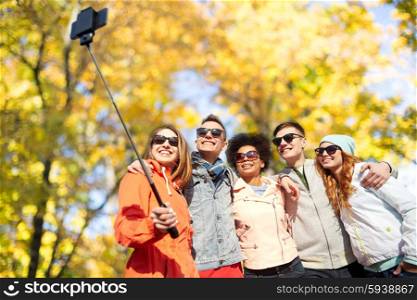 tourism, travel, people, season and technology concept - group of smiling teenage friends taking selfie with smartphone and monopod over autumn park background