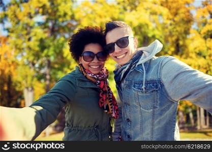 tourism, travel, people, season and technology concept - happy teenage international couple taking selfie over autumn park background