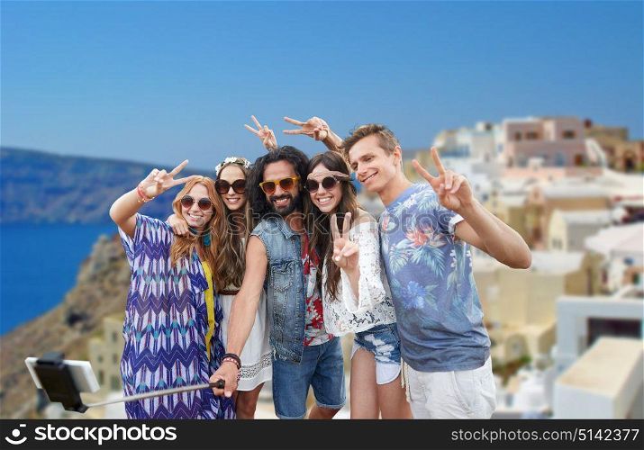 tourism, travel, technology and people concept - happy hippie friends in sunglasses taking picture by smartphone on selfie stick and showing peace gesture over oia town on santorini island background. hippie friends with smartphone on selfie stick