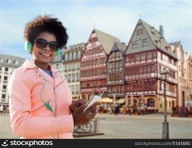 tourism, travel, technology and people concept - smiling african american young woman or teenage girl with smartphone and headphones listening to music over frankfurt am main city street background. happy young woman with smartphone and headphones
