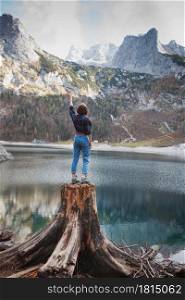 tourist girl with a backpack stands on a huge stump on the shore of a mountain lake
