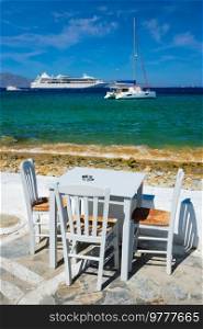 Tourist greek scene - restaurant cafe table on quay promenade with catamaran yacht and cruise liner and Aegean sea in background on beautiful summer day. Chora, Mykonos island, Greece. Catamaran yacht and cruise liner is Aegean sea. Chora, Mykonos island, Greece