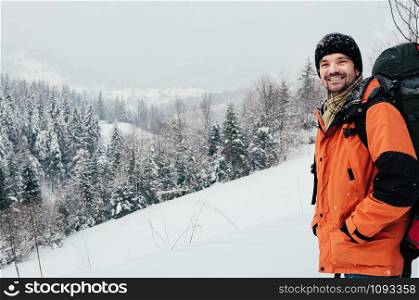 tourist male smiling into camera enjoying beautiful mountain forest landscape on snowy winter day. Orange garment, red backpack. Hiking travel extreme lifestyle concept