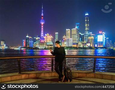 Tourist man using a smartphone near The Bund in Shanghai downtown, China in travel trip, vacation, or holidays concept in Asia. Skyscraper and high-rise buildings at night.