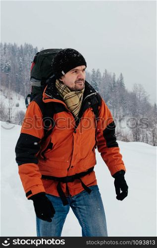 Tourist man walking through snow on winter day, mountain forest landscape. Blue jeans, orange garment, red backpack. Hiking travel extreme concept. Selective focus