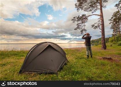 Tourist near camp tent on lake shore, looking through binoculars