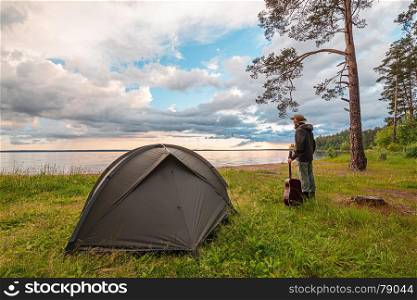 Tourist with guitar standing near camp tent on lake shore, enjoying the view at sunset. Unity with nature.