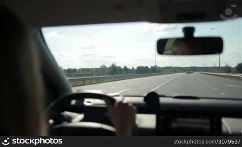 Tourist woman driving car on motorway during summer road trip. View from auto&acute;s interior. Female driver controlling vehicle on high speed on curvy asphalt road over beautiful rural scene background.