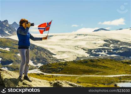 Tourist woman enjoy mountains landscape, holding norwegian flag and photo camera. National tourist scenic route 55 Sognefjellet, Norway. Tourist with camera and flag in Norway mountains