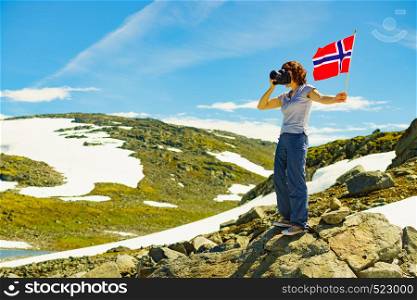 Tourist woman holding norwegian flag and taking photo with camera, snowy mountains landscape, summertime. National tourist route Aurlandsfjellet.. Tourist holds camera and norwegian flag in mountains