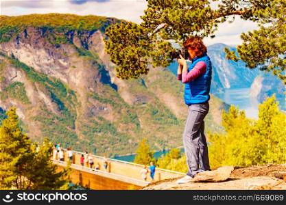 Tourist woman over Stegastein lookout taking with camera travel picture, enjoying fjord mountains landscape. National tourist scenic route Aurlandsfjellet in Norway. Tourist on nature take travel picture, Norway