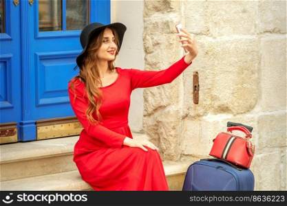 Tourist young caucasian woman in a red long dress and black hat with suitcase takes a selfie sitting on the stairs at the door outdoors. Tourist young caucasian woman in a red long dress and black hat with suitcase takes a selfie sitting on the stairs at the door outdoors.