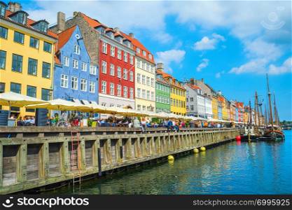 Tourists on Nyhavn embankment with cafes and restaurants in bright sunny day, boats sailing by canal, Copenhagen, Denmark