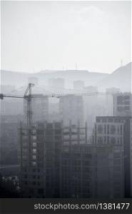 Tower crane and urban buildings. Industrial landscape with mountains background and fog.
