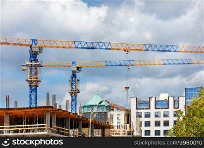 Tower cranes with a team of builders are erecting a new residential building using a monolithic frame method against a cloudy sky.. Construction site with tower cranes and the construction of a residential building against the backdrop of a cloudy sky.