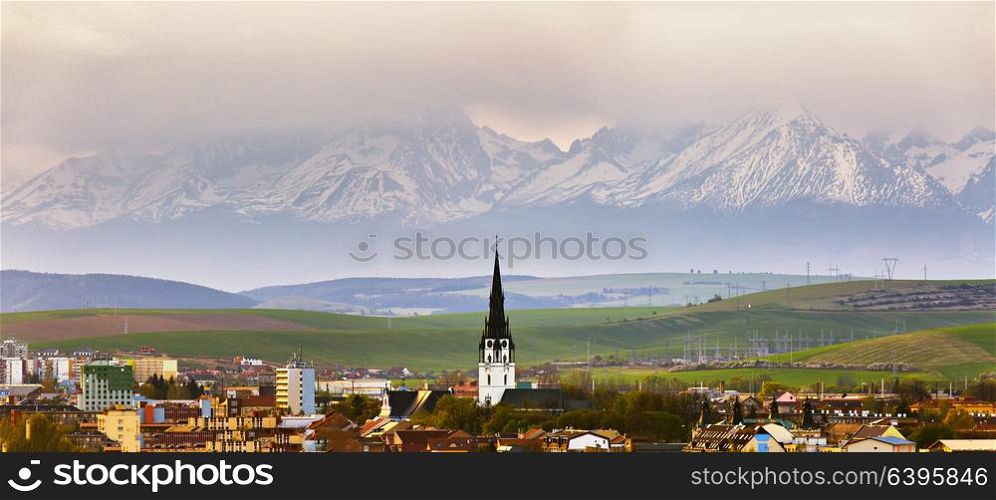 Town and peak of cathedral tower behind snow-caped mountain range. Church and spring green cultivated fields.