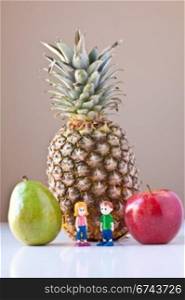 Toy girl and boy standing in front of pineapple, red apple and green pear on white with taupe brown background. The concepts depicted in this image are nutrition, good food choices, balanced diet and good for you.