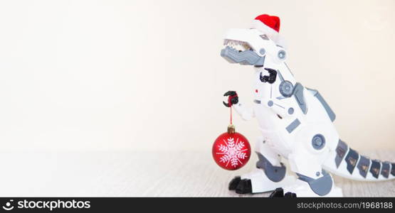 toy robot dinosaur in a santa hat and with a fir ball on a beige background. copy space.