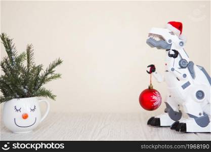 toy robot dinosaur in a Santa hat and with a fir ball and a snowman mug with fir branches on a beige background. copy space.